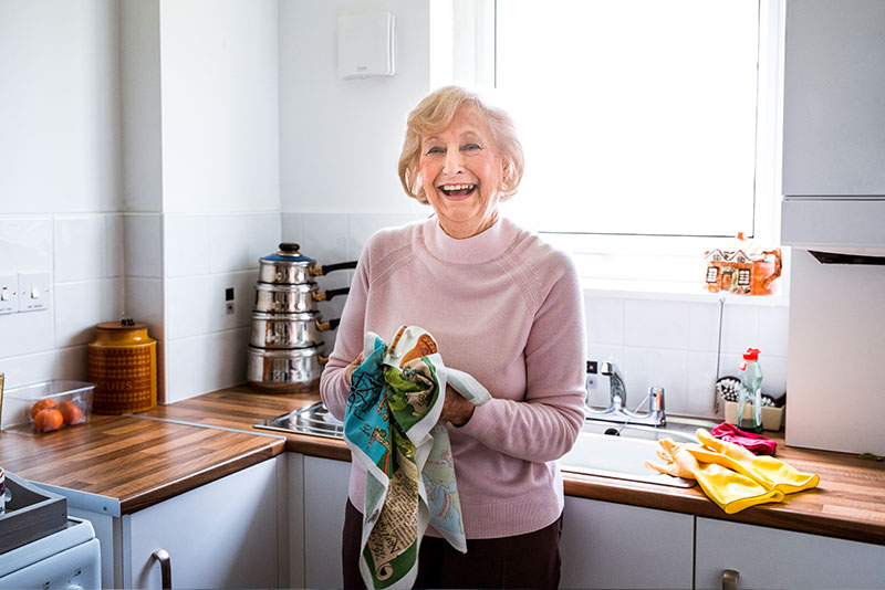 A woman experiencing enhanced independent living for someone with dementia smiles as she dries dishes in the kitchen.
