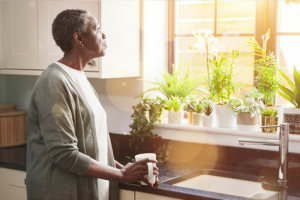 A woman gazes out the window while holding a cup of coffee, pondering whether or not she may be displaying the signs of caregiver burden.