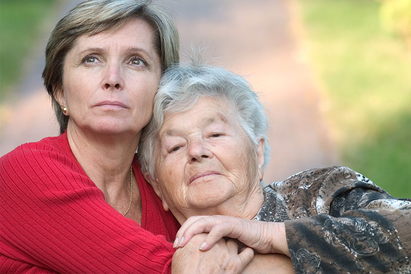 A woman whose mom has Alzheimer’s hugs her while gazing thoughtfully into the distance.