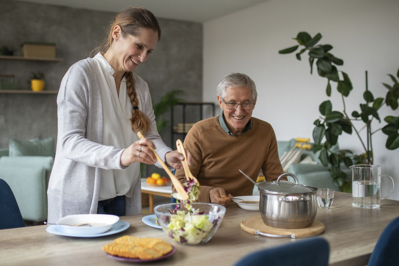 An older man experiencing senior nutrition challenges smiles as his caregiver prepares a meal for him.