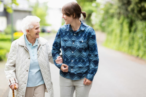 A caregiver helps with preventing falls at home by offering her arm to steady an older woman walking with a cane.