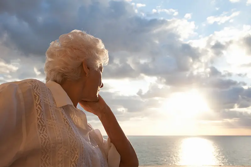 An older woman in need of summer grief support for seniors stares out at the water during a sunset.